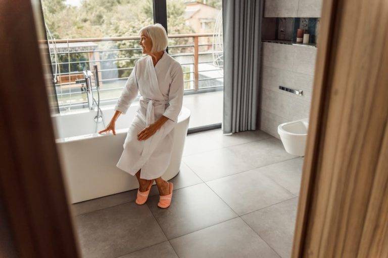 elderly woman sat on the edge of a bath tub in a dressing gown